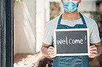 Closeup of one mixed race man wearing a face mask and holding a chalkboard with the word "welcome" written on in a cafe or store. Hands of guy holding sign to greet customers at the entrance of a shop open during covid pandemic