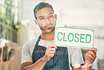 Portrait of one young hispanic man holding a "closed" sign at a window on display in a cafe or store. Mixed race guy showing the closure of his small business or shop due to bankruptcy and recession
