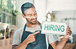 One young hispanic man pointing to a "hiring" sign at a window on display in a cafe or store. Happy mixed race guy advertising job opportunity while recruiting new staff for his startup shop