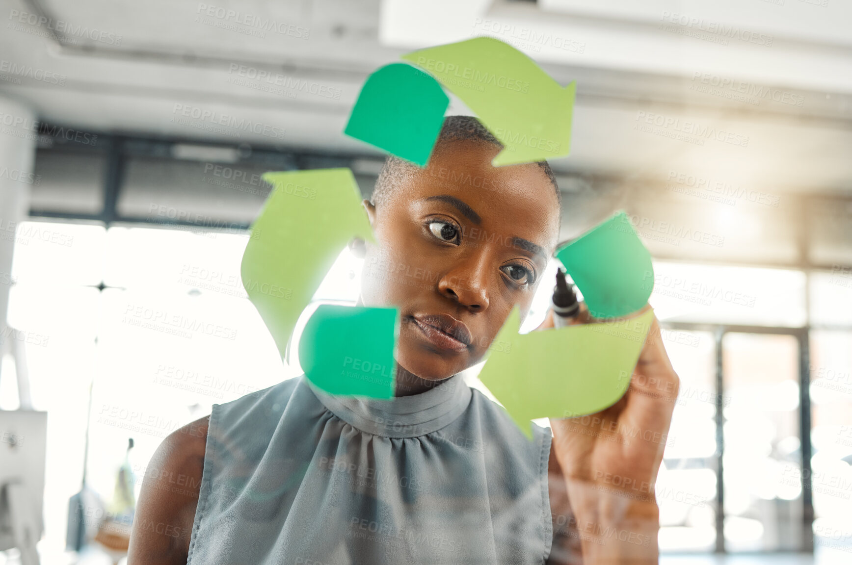 Buy stock photo Young focused african american businesswoman drawing a recycle symbol on a glass window in an office at work. One serious black female businessperson designing a sign for awareness to recycling
