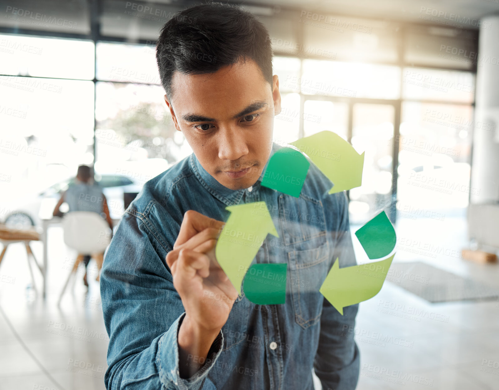 Buy stock photo Young focused mixed race businessman drawing a recycle symbol on a glass window in an office at work. One serious hispanic businessperson designing a sign for awareness to recycling