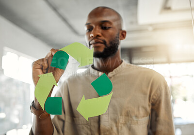 Buy stock photo Young focused african american businessman drawing a recycle symbol on a glass window in an office at work. One serious male businessperson designing a sign for awareness to recycling