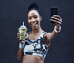 One happy young mixed race woman drinking a healthy green detox smoothie and taking selfies on phone while exercising against dark background. Female athlete sipping on fresh nutritious fruit juice in plastic cup while taking photos for social media