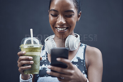Closeup of one happy young mixed race woman drinking a healthy green detox smoothie and texting on phone while exercising against dark background. Female athlete sipping on fresh nutritious fruit juice in plastic cup to cleanse while browsing social media