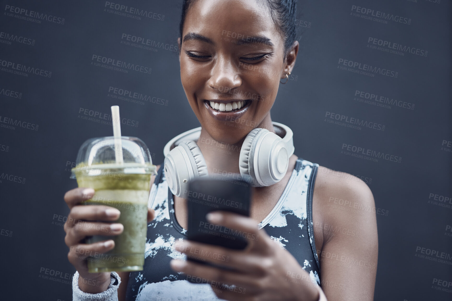 Buy stock photo Closeup of one happy young mixed race woman drinking a healthy green detox smoothie and texting on phone while exercising against dark background. Female athlete sipping on fresh nutritious fruit juice in plastic cup to cleanse while browsing social media