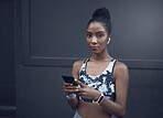 Portrait of one young mixed race woman listening to music with wireless earphones from a cellphone while on a break from exercise outdoors against a dark background. Focused female athlete texting, using fitness apps online and browsing social media