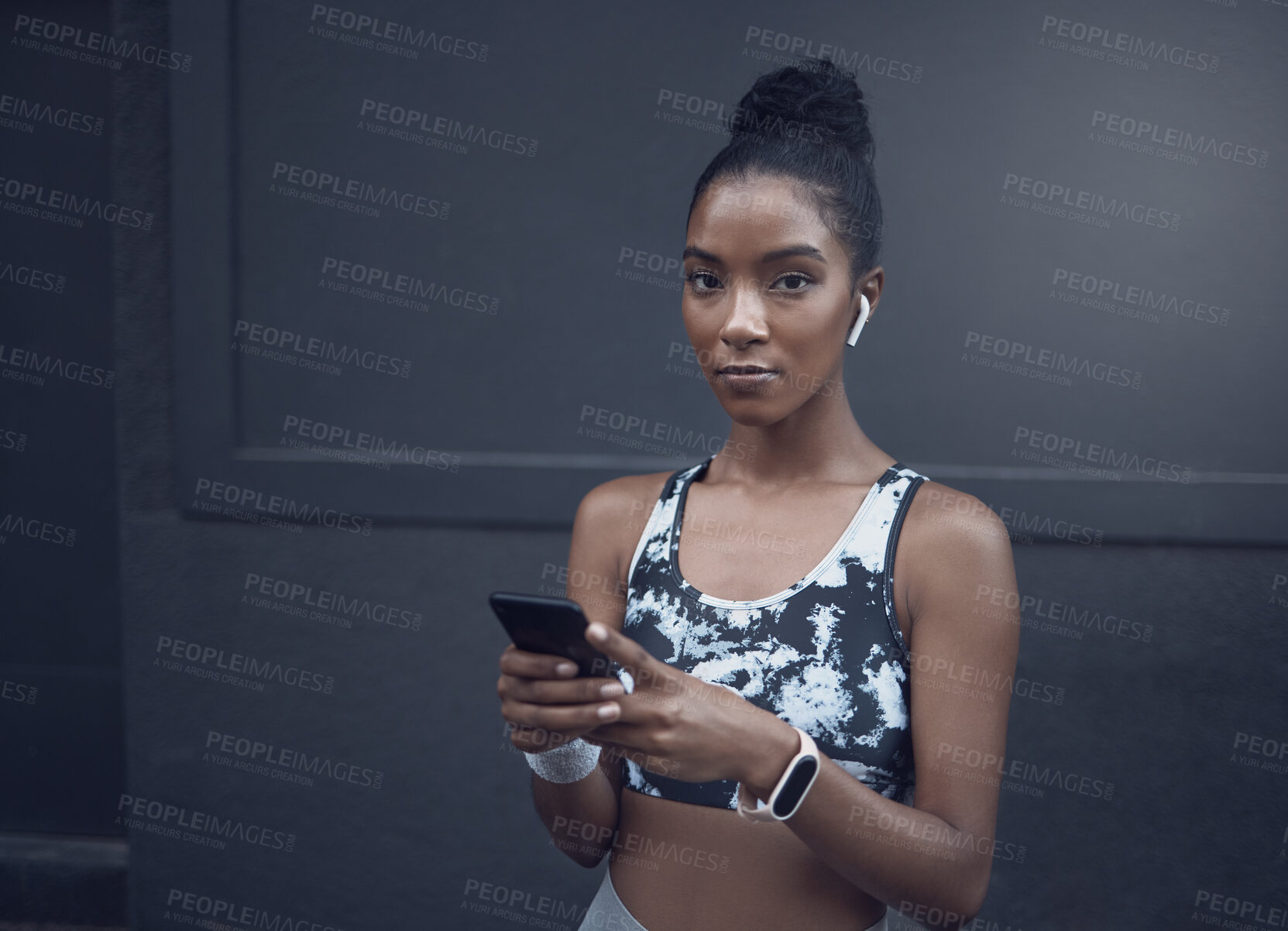 Buy stock photo Portrait of one young mixed race woman listening to music with wireless earphones from a cellphone while on a break from exercise outdoors against a dark background. Focused female athlete texting, using fitness apps online and browsing social media
