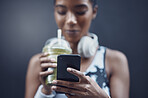 Closeup of one mixed race woman drinking a healthy green detox smoothie and texting on phone while exercising against dark background. Female athlete sipping on fresh nutritious fruit juice in plastic cup to cleanse while browsing social media