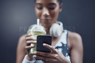 Buy stock photo Closeup of one mixed race woman drinking a healthy green detox smoothie and texting on phone while exercising against dark background. Female athlete sipping on fresh nutritious fruit juice in plastic cup to cleanse while browsing social media