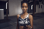 One fit young mixed race woman listening to music with wireless earphones from a cellphone while taking a break from exercise outdoors. Focused female athlete texting and using fitness apps online while browsing social media and watching workout tutorials