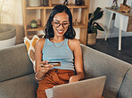 Smiling mixed race woman using credit card for ecommerce on laptop at home. Happy hispanic sitting alone on living room sofa and using technology for ebanking. Relaxing, ordering and buying online