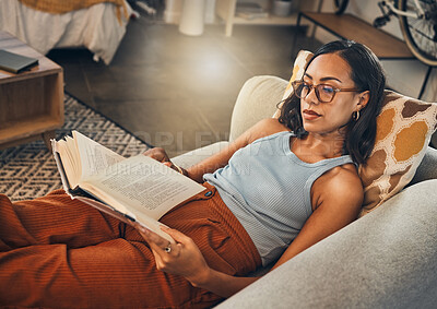 Serious mixed race woman reading a book in living room at home. Beautiful hispanic lying down on lounge sofa alone and enjoying a novel. Relaxed woman wearing glasses to read fiction story on weekend