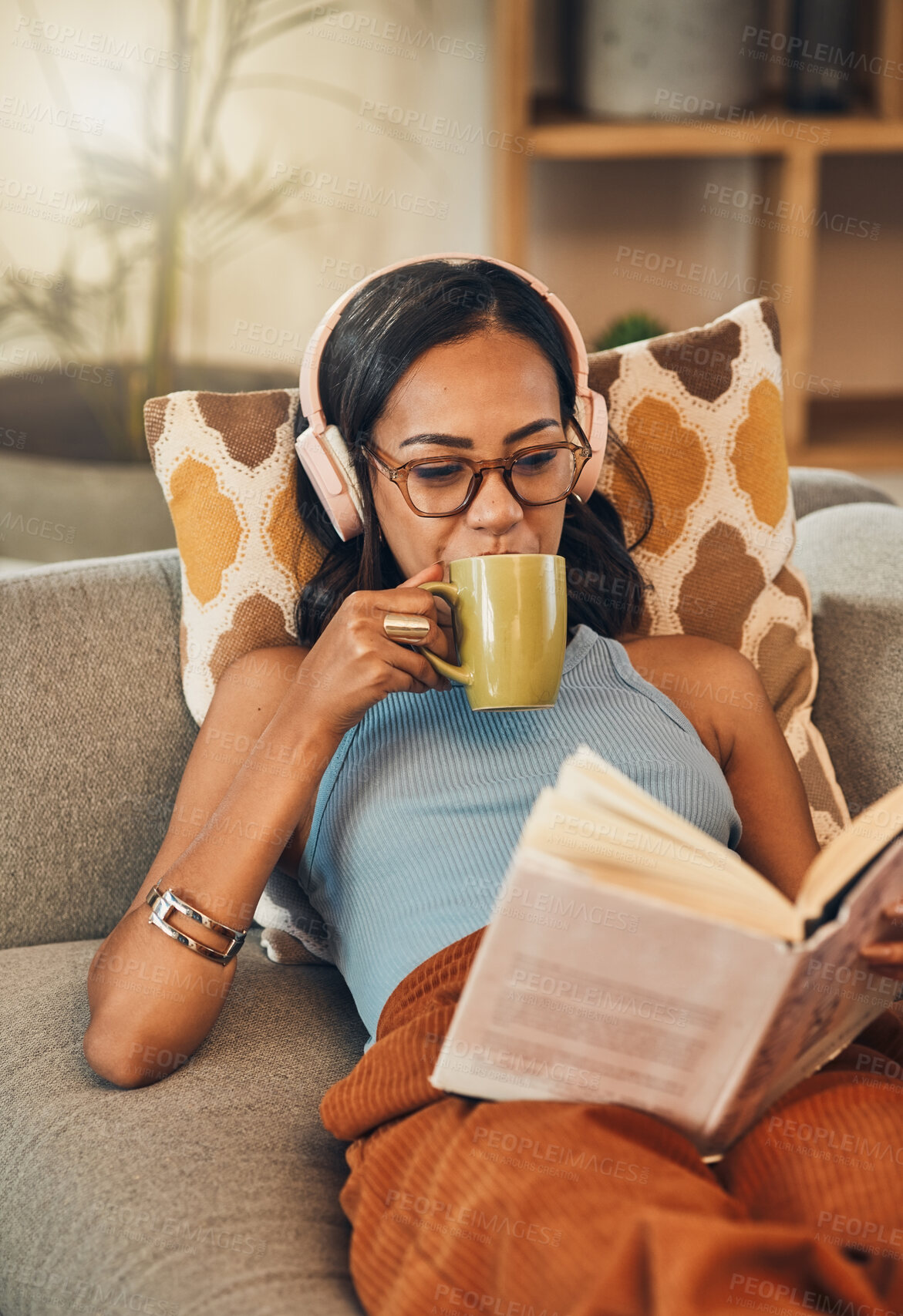 Buy stock photo Beautiful mixed race woman reading book and drinking coffee while listening to music on headphones in living room at home. Hispanic lying down on lounge sofa alone and enjoying novel. Feeling relaxed