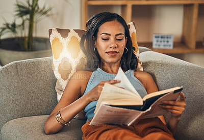 Serious mixed race woman reading a book in living room at home. Beautiful hispanic lying down on lounge sofa alone and enjoying a novel. Relaxed woman wearing glasses to read fiction story on weekend