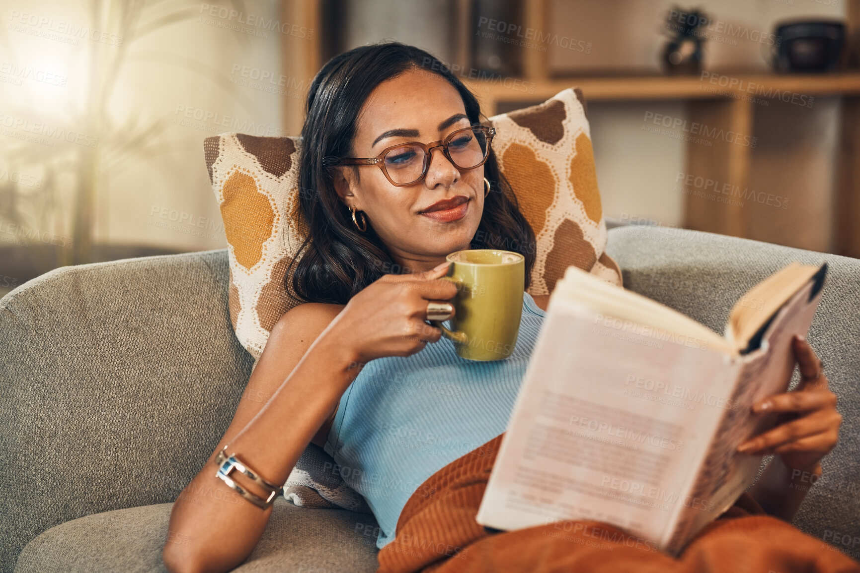 Buy stock photo Beautiful mixed race woman reading book and drinking coffee in living room at home. Hispanic lying down on lounge sofa alone and enjoying novel. Feeling relaxed on weekend with fiction story and tea
