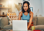 Beautiful mixed race woman using laptop for blogging while listening to music on headphones in living room at home. Hispanic entrepreneur sitting alone on lounge sofa, typing blog post on technology