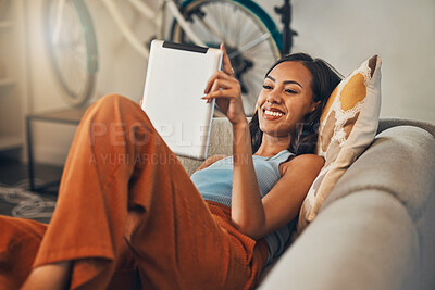 Smiling mixed race woman browsing internet on digital tablet at home. Happy hispanic lying down on living room sofa alone and using technology. Relaxed woman scrolling and searching online on weekend