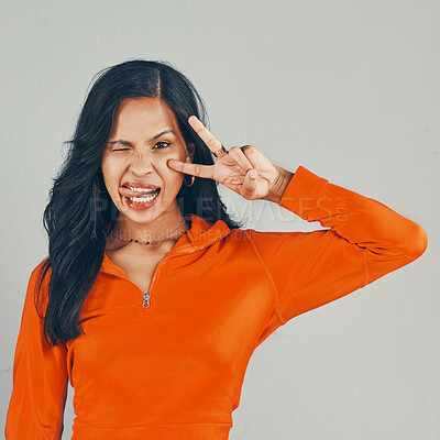 Buy stock photo Portrait of mixed race woman isolated against grey studio background with copyspace. Young playful hispanic standing alone, making peace sign and symbol with hand gesture and winking with tongue out