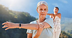 Senior woman stretching her arms while exercising outdoors on a sunny day. Mature people practicing yoga together in nature staying healthy and active during retirement. Wearing smart watch
