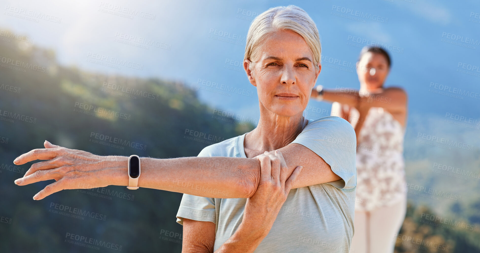 Buy stock photo Senior woman stretching her arms while exercising outdoors on a sunny day. Mature people practicing yoga together in nature staying healthy and active during retirement. Wearing smart watch