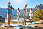Three senior people meditating with joined hands and closed eyes breathing deeply while standing on yoga mats. Multiethnic class of mature people doing yoga together in nature on a sunny day