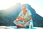Active senior woman using her smart phone while sitting on her yoga mat with scenic mountain view in the background. Woman sending text or using mobile app while practicing yoga