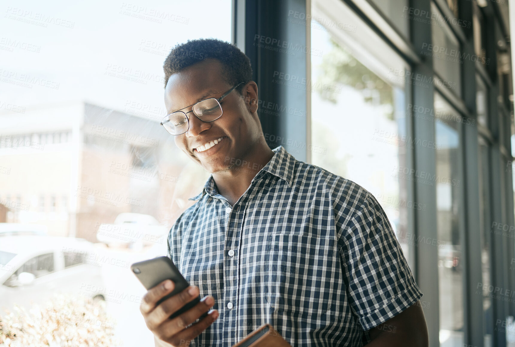 Buy stock photo Happy african american business man entrepreneur leaning against a window in an office while reading or sending text on smartphone. Smiling entrepreneur chatting online or getting good news via app