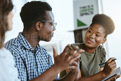 Three young happy businesspeople having a meeting while sitting at a table and taking notes in a notebook at work. Business professionals talking and planning in an office together. African american woman discussing a business strategy with coworkers