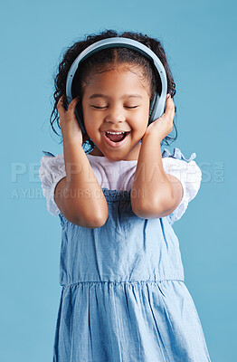 Adorable little hispanic girl standing with her eyes closed and looking happy while wearing wireless headphones and enjoying her favourite song against a blue studio background