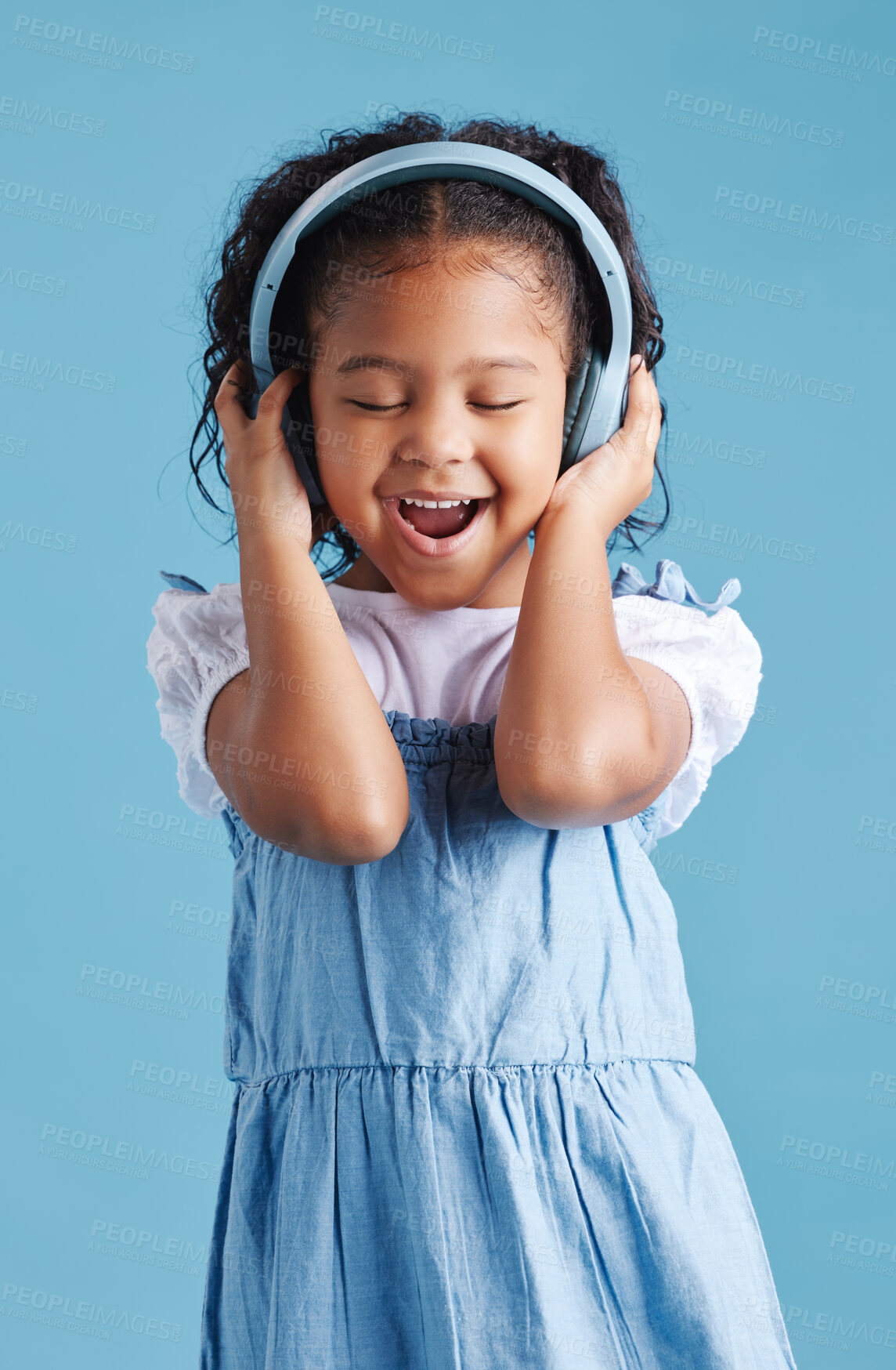 Buy stock photo Adorable little hispanic girl standing with her eyes closed and looking happy while wearing wireless headphones and enjoying her favourite song against a blue studio background