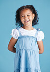 Happy smiling little girl standing with her hands behind her back against blue studio background. Cheerful mixed race kid in casual denim dress and white tshirt