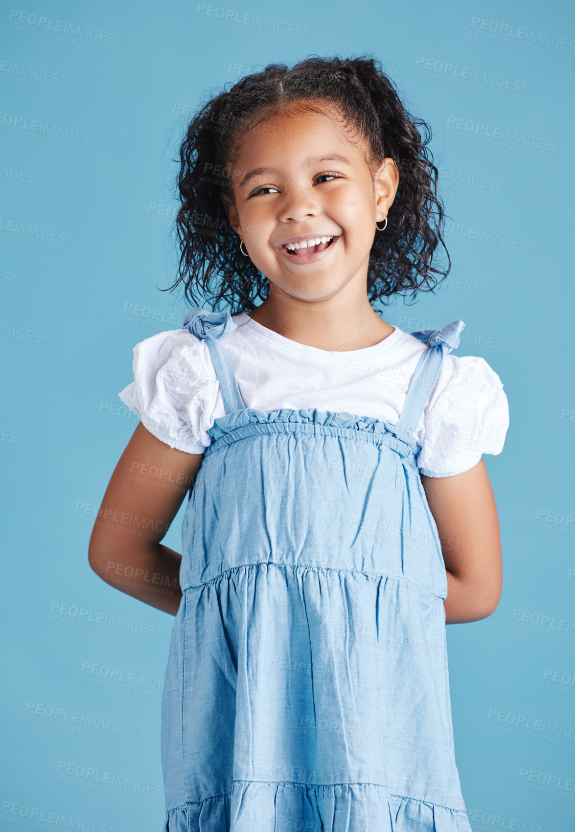 Buy stock photo Happy smiling little girl standing with her hands behind her back against blue studio background. Cheerful mixed race kid in casual denim dress and white tshirt