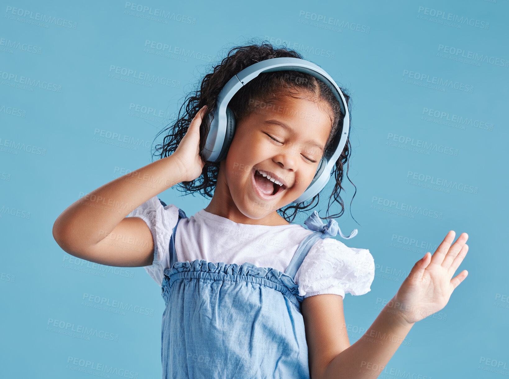 Buy stock photo Adorable little hispanic girl standing with her eyes closed and looking happy while wearing headphones and listening to music, dancing to her favourite song against a blue studio background