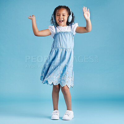 Full length of an adorable little hispanic girl dancing with her hands up looking happy while listening to music with wireless headphones enjoying her favourite song against a blue studio background