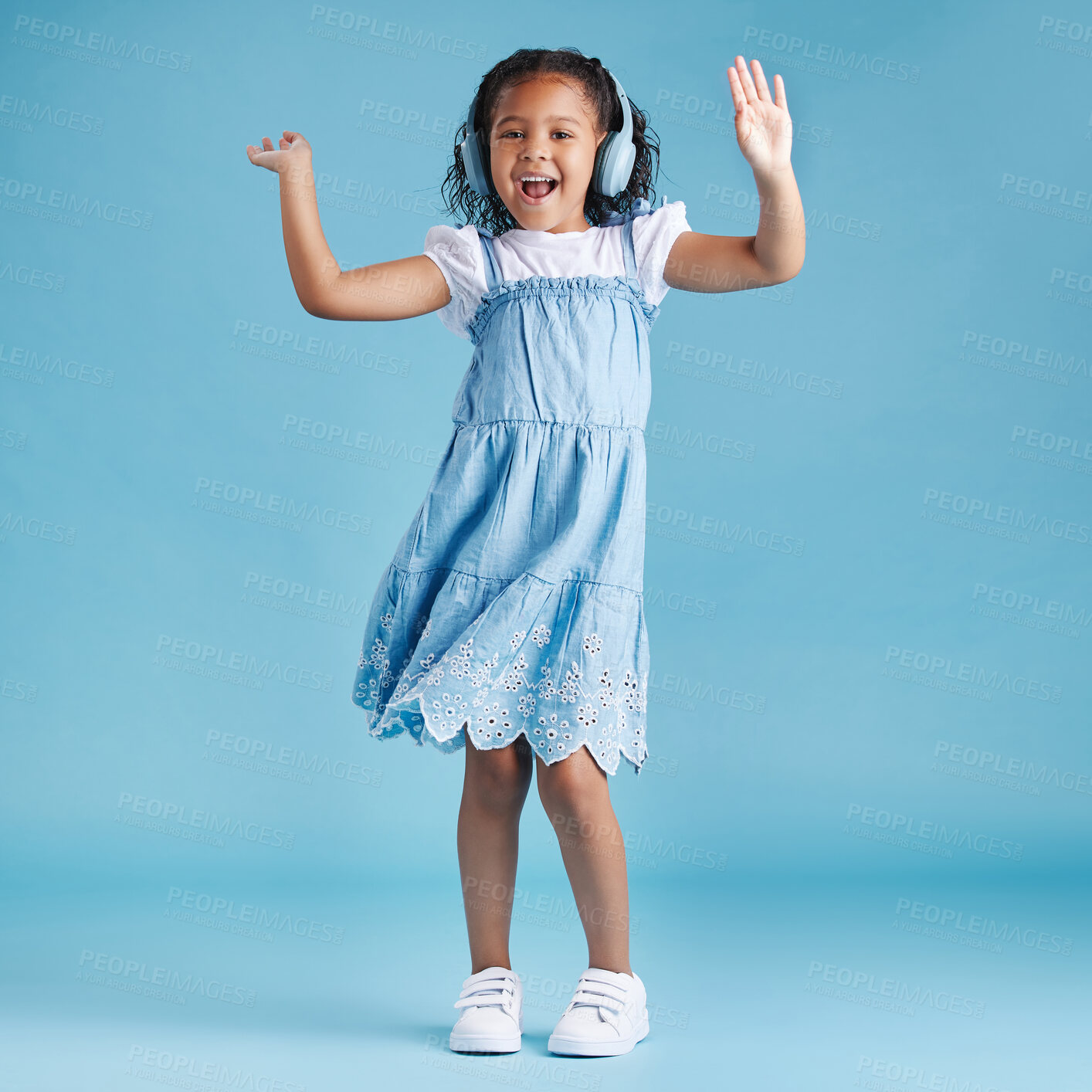 Buy stock photo Full length of an adorable little hispanic girl dancing with her hands up looking happy while listening to music with wireless headphones enjoying her favourite song against a blue studio background