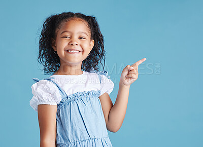 Portrait of happy smiling little girl pointing her finger at copy space to the right against blue studio background. Cheerful mixed race kid in casual clothes