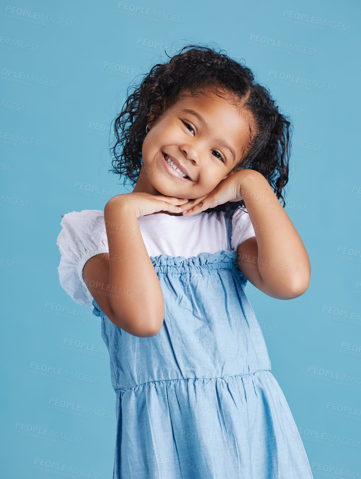 Buy stock photo Portrait of happy smiling little girl posing with her hands under her face showing a healthy dental smile against blue studio background. Cute mixed race kid in casual clothes