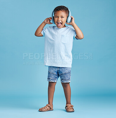 Full length of an excited little boy listening to music with wireless headphones and wearing casual clothes against a blue studio background