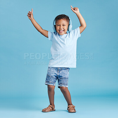 Full length of a funny little boy dancing while listening to music with wireless headphones and wearing casual clothes against a blue studio background