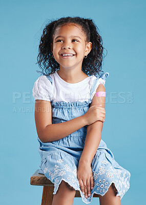 Happy vaccinated little girl kid sitting on chair showing arm with adhesive bandage after vaccine injection standing against a blue studio background. Advertising vaccination against coronavirus. Child immunisation