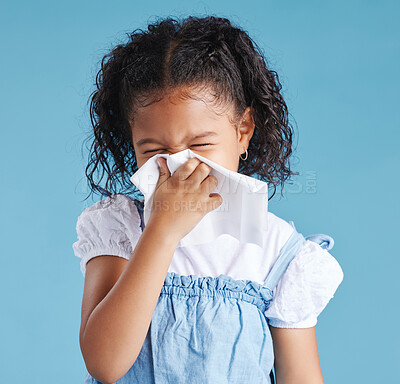 Little hispanic girl blowing her nose with tissue against blue studio background. Child feeling sick with flu virus or infection and suffering with snotty nose