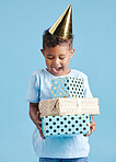Cheerful little boy in casual clothes and party hat while standing against blue background and holding birthday presents. Cute kid excited about opening his gifts