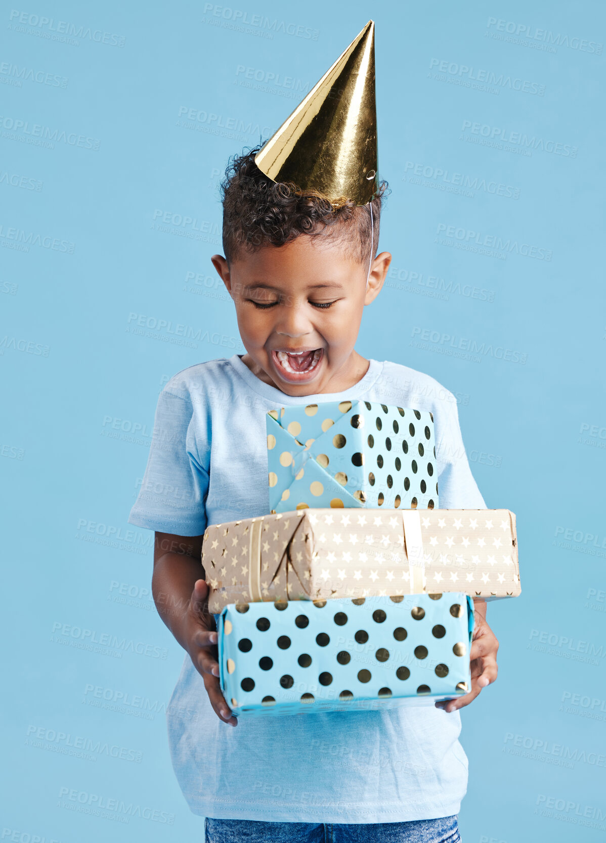 Buy stock photo Cheerful little boy in casual clothes and party hat while standing against blue background and holding birthday presents. Cute kid excited about opening his gifts