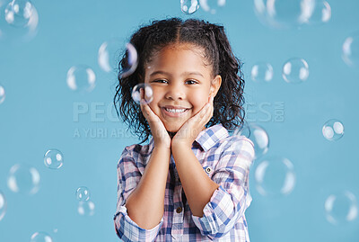 Portrait of happy smiling little girl posing with her hands on her face with bubbles falling around her against blue studio background. Cute mixed race kid in casual clothes