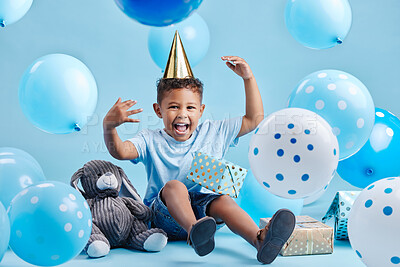Excited little boy celebrating his birthday with balloons and gifts on a blue studio background. Cute kid wearing a party hat and sitting with presents and a stuffed bunny