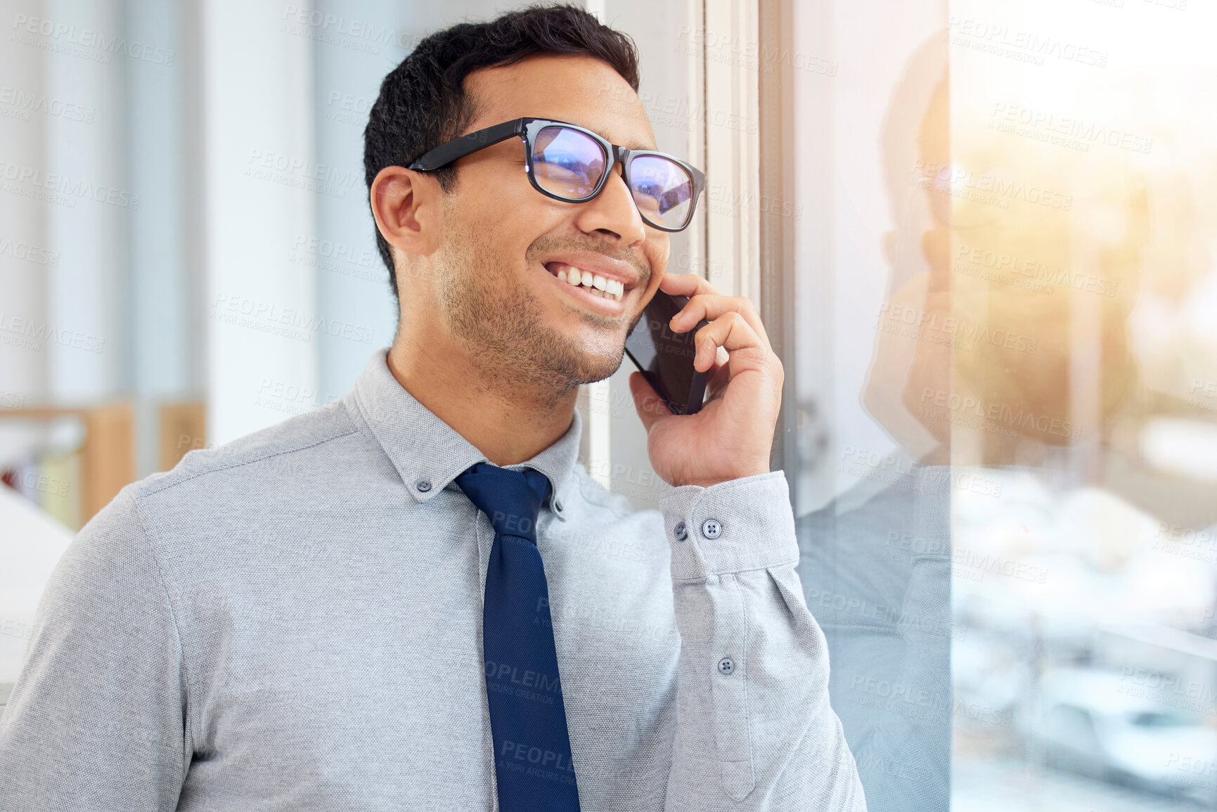 Buy stock photo Young content mixed race businessman on a call using a phone and  standing in an office at work. One hispanic male businessperson smiling while talking on the phone standing and thinking at a window in an office