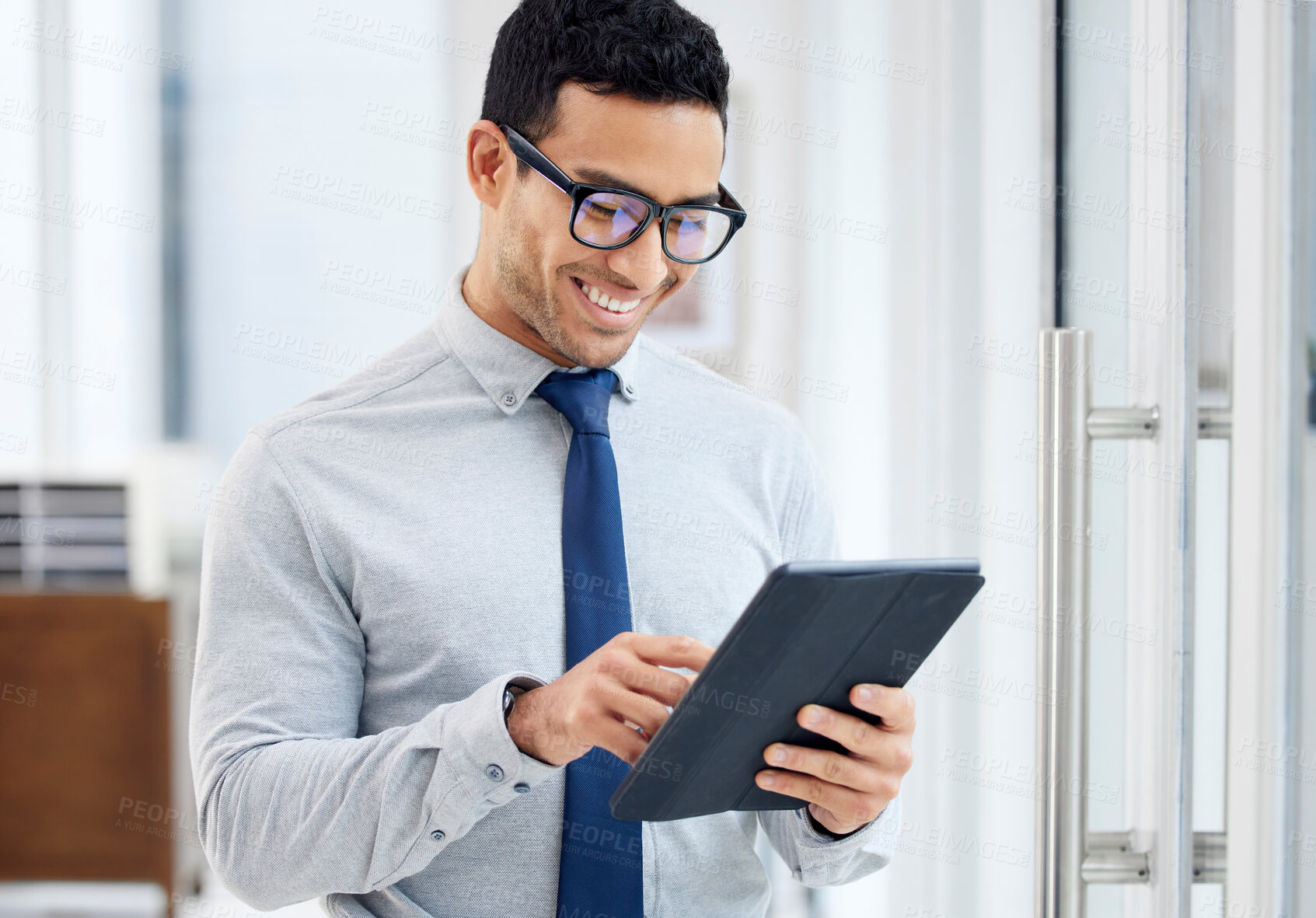 Buy stock photo Happy mixed race businessman holding and using a digital tablet standing in an office at work. One content hispanic male businessperson smiling while typing an email on a digital tablet at work
