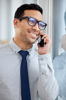 Young happy mixed race businessman on a call using a phone and standing in an office at work. One hispanic male businessperson wearing glasses smiling while talking on the phone standing and thinking at a window in an office