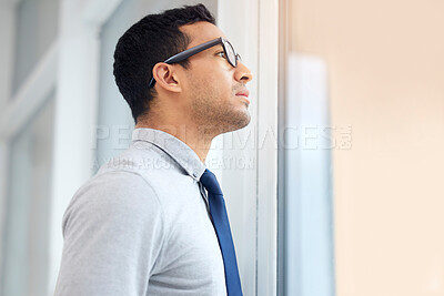 Buy stock photo Serious mixed race businessman looking out of a window standing alone in an office at work. One hispanic standing and thinking while wearing glasses at work