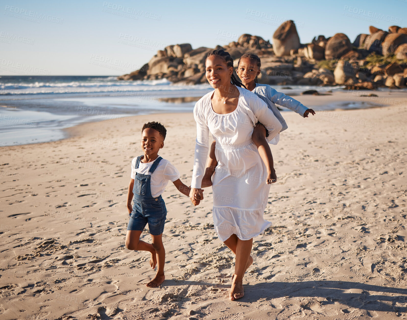 Buy stock photo Portrait of a beautiful young african american mother at the beach with her two children. Little girl sitting on her moms back while her brother holds her hands as they walk along the beach